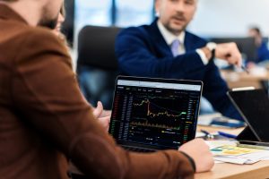A person holding a laptop displaying a cryptocurrency trading chart during a business meeting, with a colleague in a suit discussing data in the background.