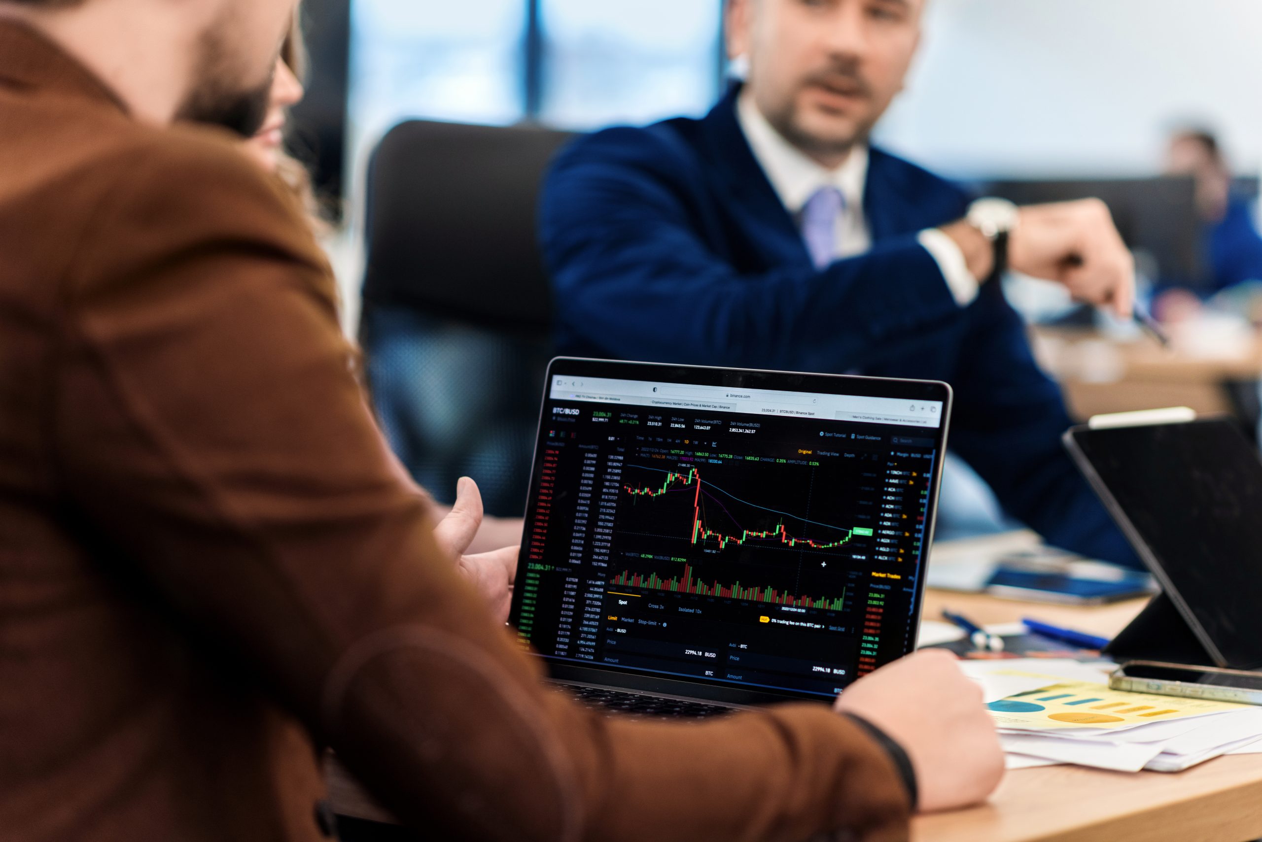 A person holding a laptop displaying a cryptocurrency trading chart during a business meeting, with a colleague in a suit discussing data in the background.