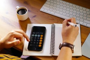 A person is working at a wooden desk, using a smartphone calculator to perform calculations while writing in a spiral notebook. A cup of black coffee, a white keyboard, and part of a laptop are visible on the desk, indicating a workspace setup. The individual is wearing a wristwatch and holding a pen, suggesting focus and engagement in financial or business-related tasks.