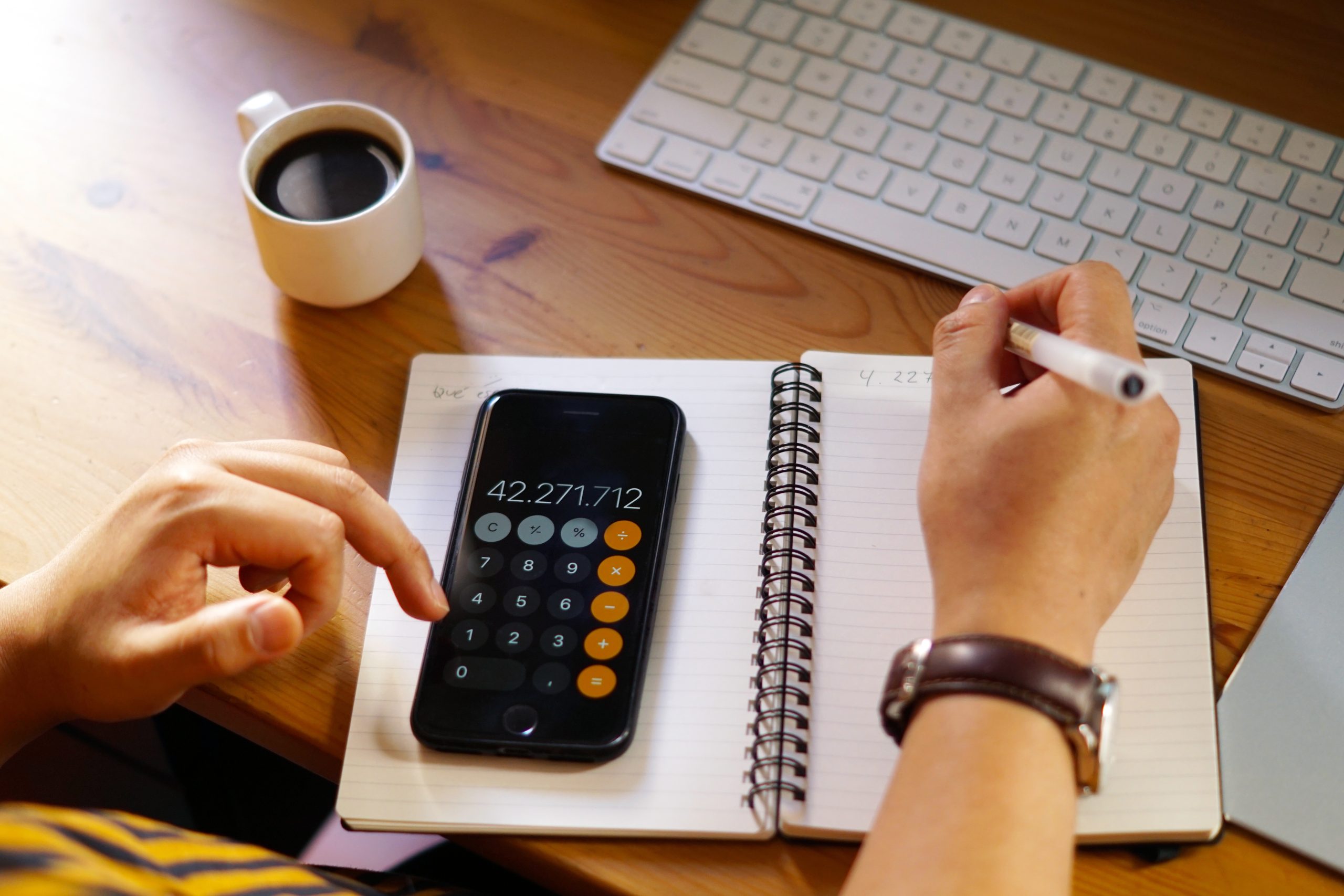 A person is working at a wooden desk, using a smartphone calculator to perform calculations while writing in a spiral notebook. A cup of black coffee, a white keyboard, and part of a laptop are visible on the desk, indicating a workspace setup. The individual is wearing a wristwatch and holding a pen, suggesting focus and engagement in financial or business-related tasks.
