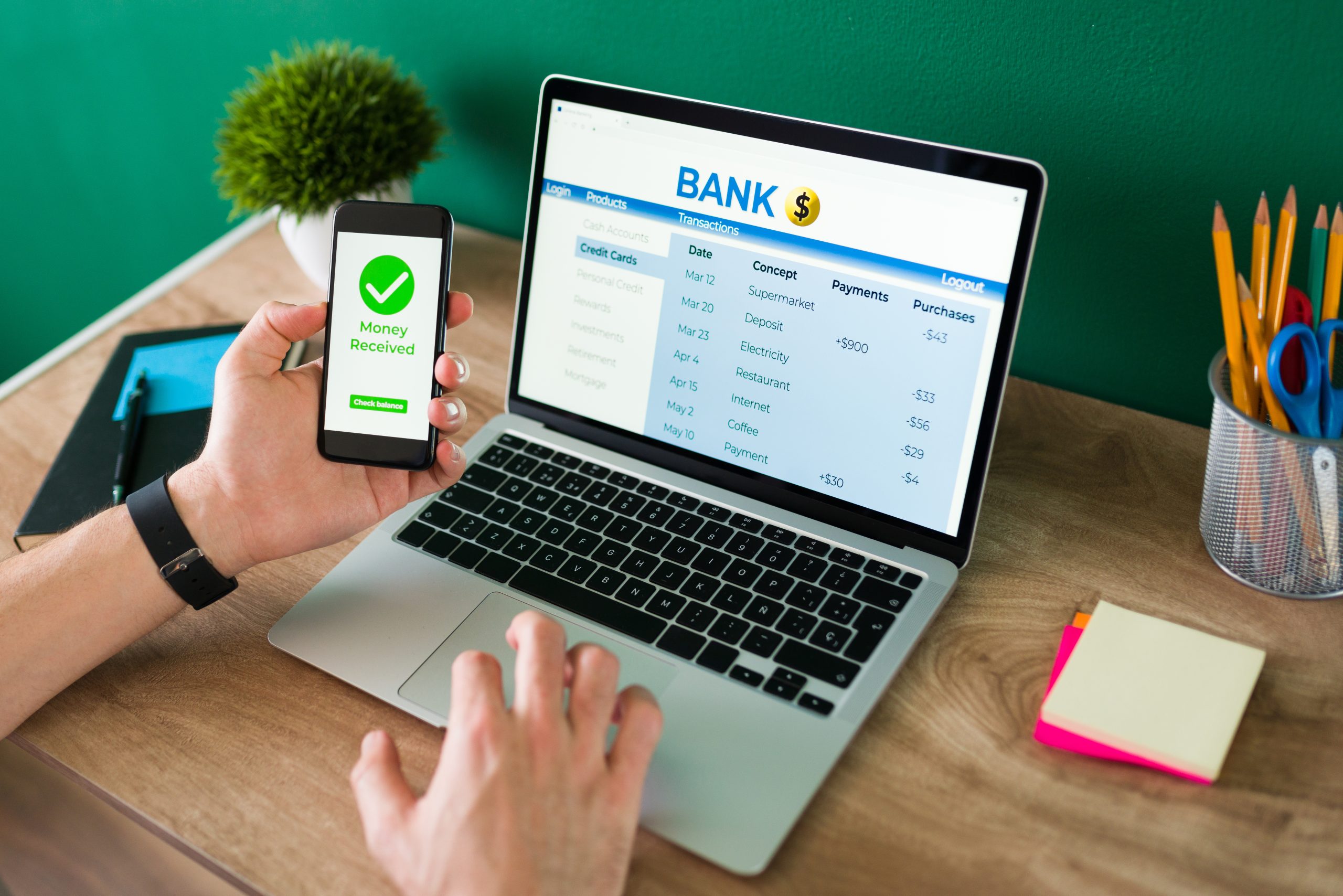 A person holding a smartphone displaying a "Money Received" confirmation while working on a laptop showing an online banking account with transaction details, sitting at a desk with office supplies.