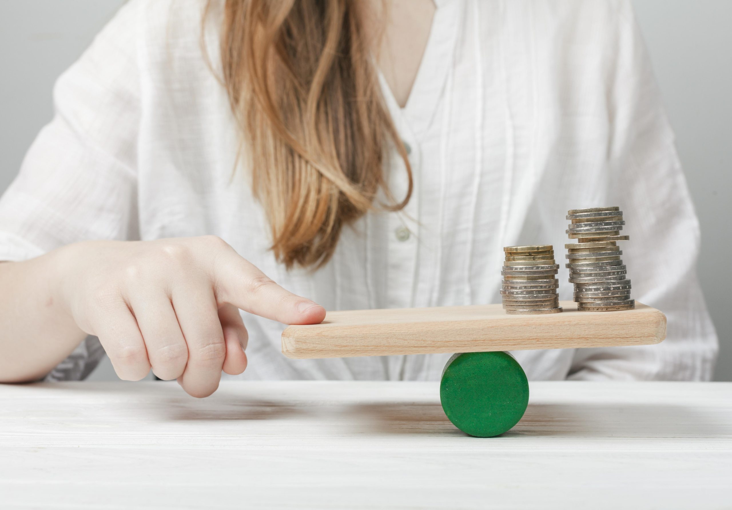 A woman in a white blouse is seen balancing a wooden plank on a green cylindrical support while pressing one end with her finger. On the other end of the plank, there are neatly stacked coins of different sizes and colors, symbolizing financial balance and stability. The background is neutral, emphasizing the concept of money management and financial equilibrium.