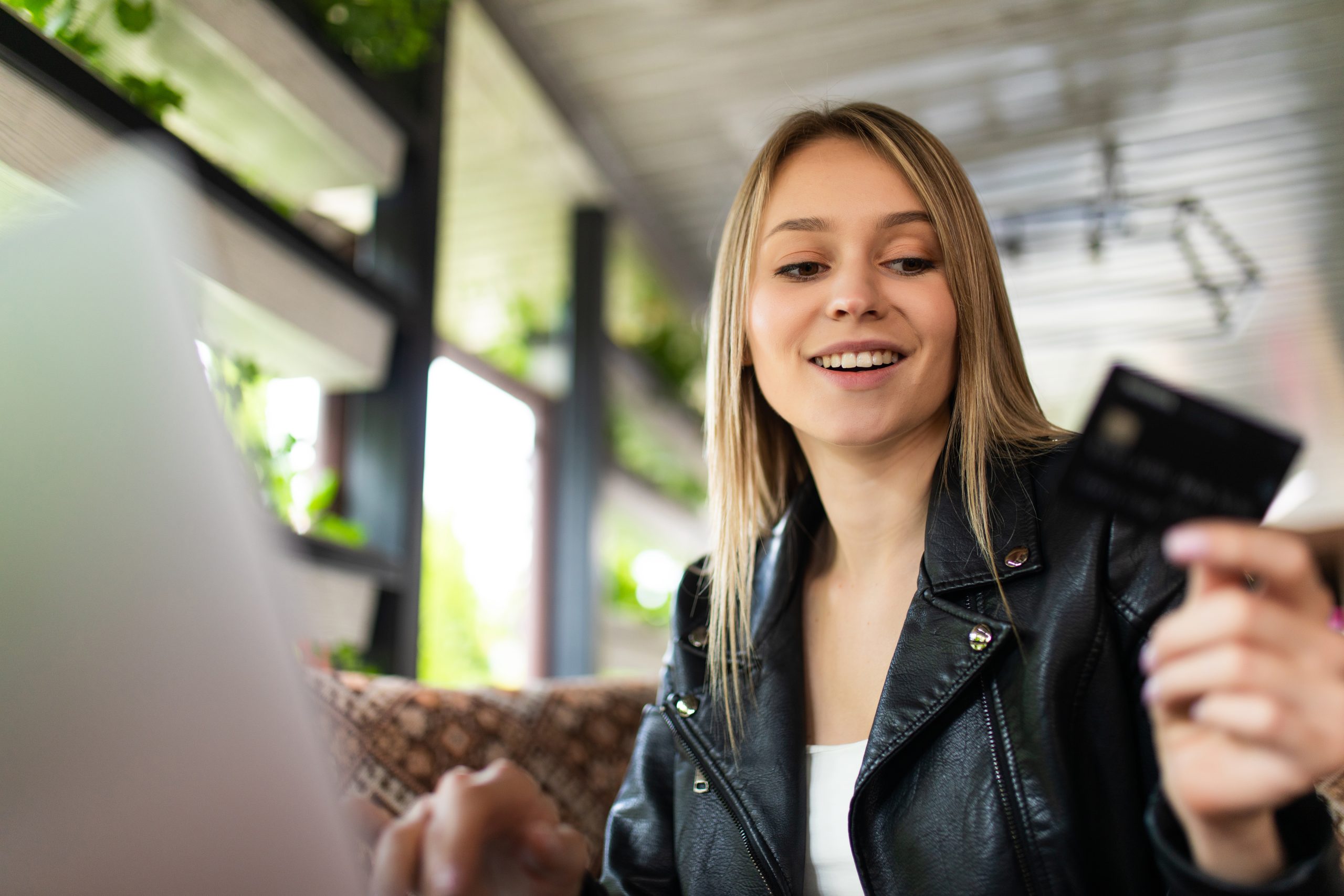A young woman wearing a black leather jacket is sitting in a modern, well-lit space with green plants, holding a credit card while using her laptop. She has a cheerful expression, suggesting she is making an online purchase or managing her finances. The setting appears to be a café or a cozy workspace, emphasizing the convenience of digital transactions.