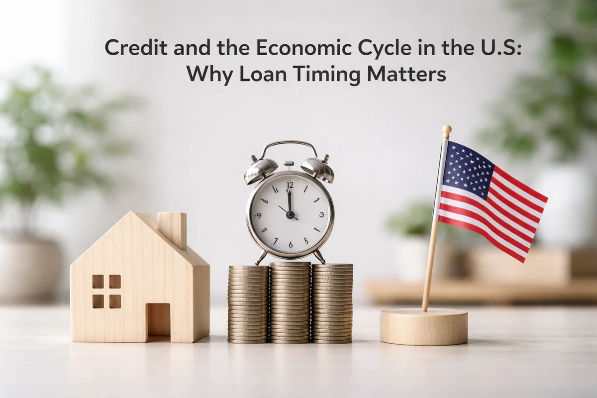 A conceptual financial image illustrating the relationship between the economic cycle and loans in the United States. On a clean surface, a small wooden house model stands beside neatly stacked coins topped with a classic alarm clock, symbolizing timing and interest rate cycles. A small U.S. flag on a wooden base reinforces the national context. The composition visually represents how economic conditions and timing influence loans, borrowing decisions, and long-term financial planning in the U.S. market.